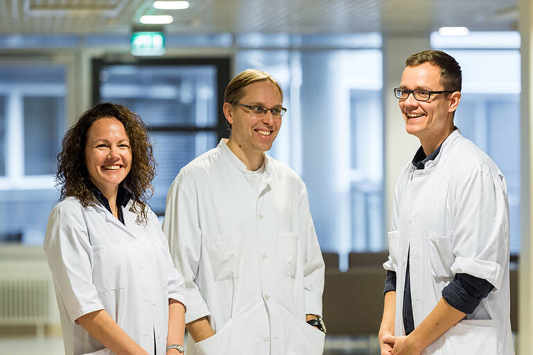Kolme tutkijaa hymyilee iloisesti valkoisissa takeissa.